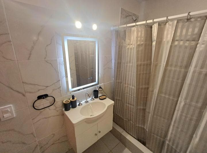 Baño con vanitory, espejo iluminado y mampara de ducha con azulejos marmoleados.