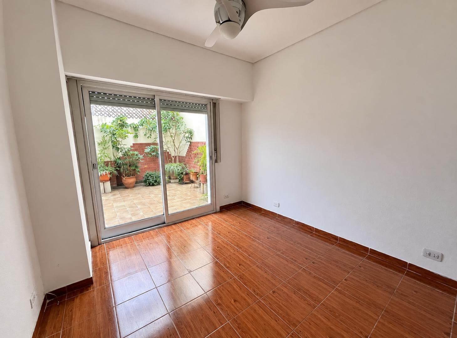Dormitorio vacío con ventilador de techo, piso de baldosas y puerta balcón al patio.