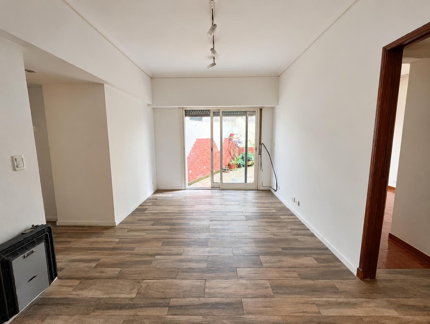 Living vacío con piso de madera, estufa y puerta ventana que da al patio.