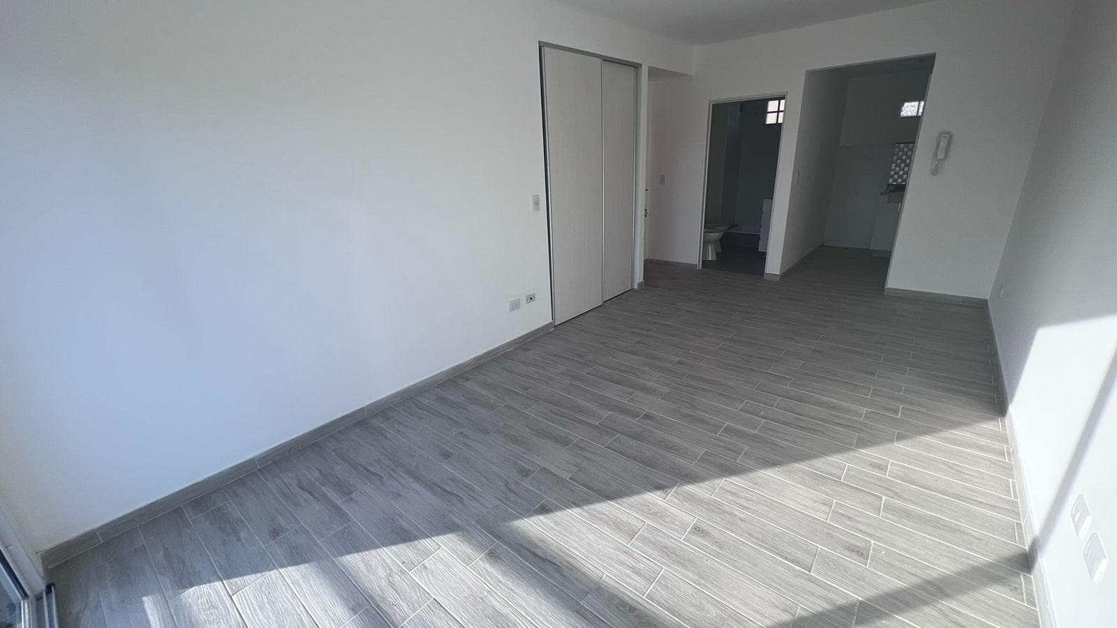 Living vacío con piso de cerámico gris, entrada al baño y la cocina.