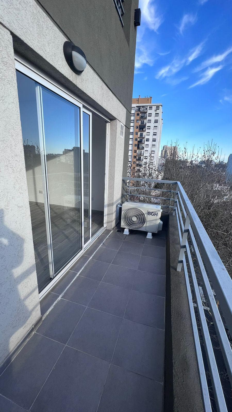 Balcón estrecho con baranda metálica, vista urbana y unidad exterior de aire acondicionado.