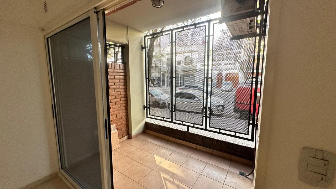 Balcón cerrado con rejas, pared de ladrillo y vista a la calle.
