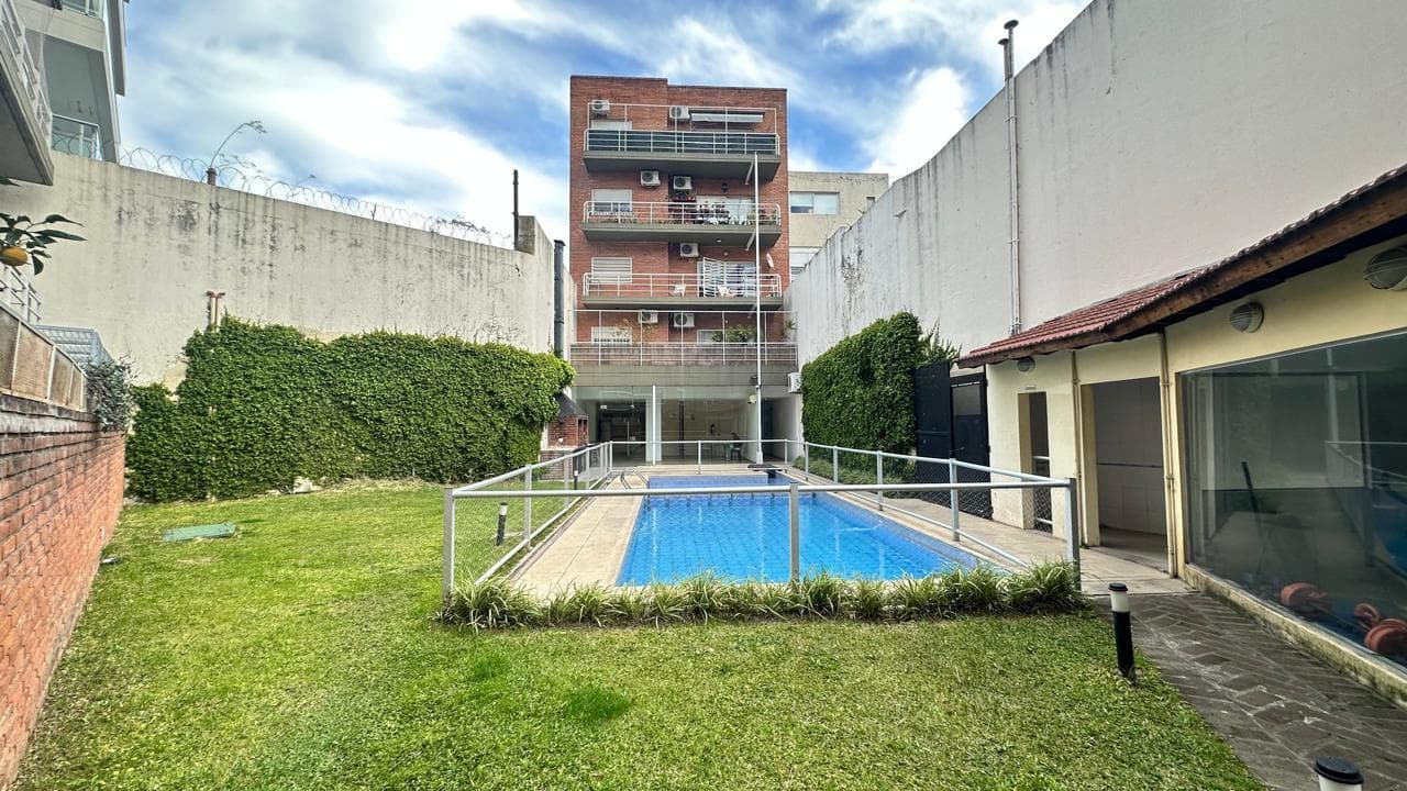 Amplio jardín con pileta y edificio de ladrillo de fondo.