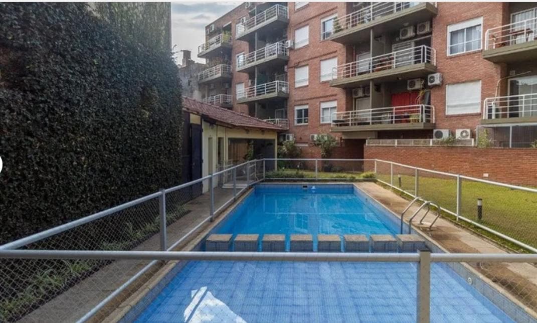Pileta exterior con cerco y edificio de ladrillo rodeado de vegetación.