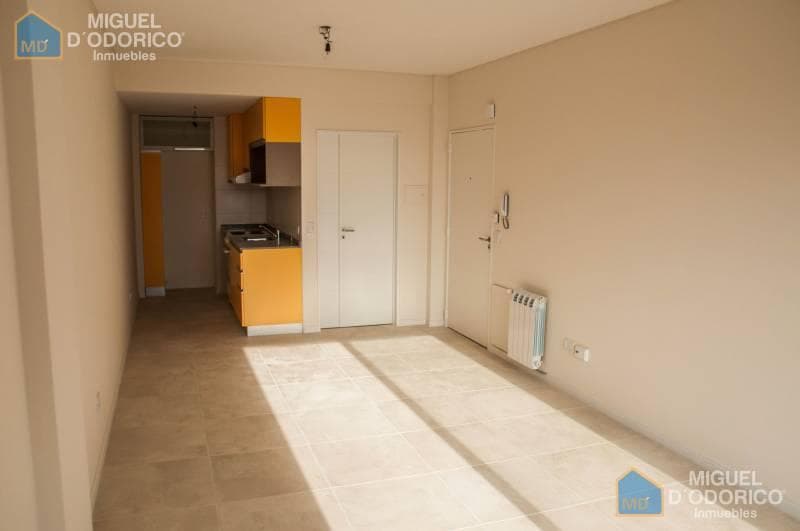 Living vacío con cocina integrada y puerta de entrada al departamento.
