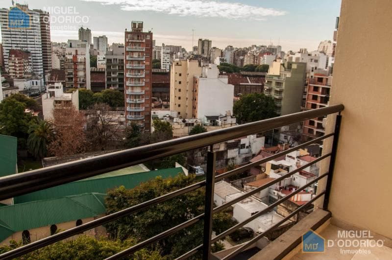 Vista de la ciudad desde balcón con baranda de metal, edificios y árboles.