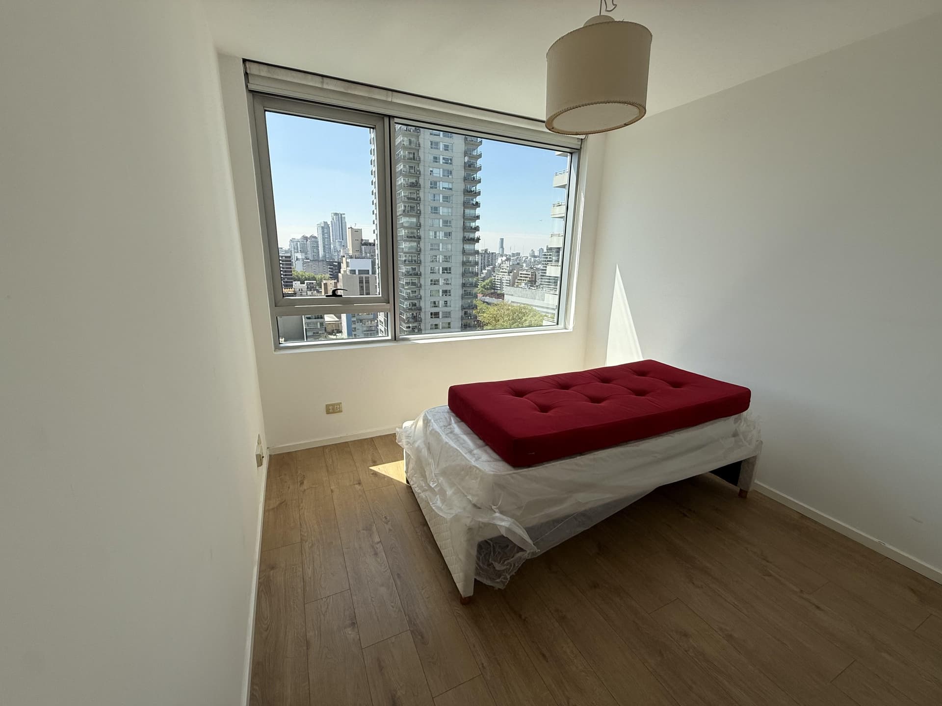 Dormitorio con colchón rojo sobre base y ventana grande con vista a la ciudad.