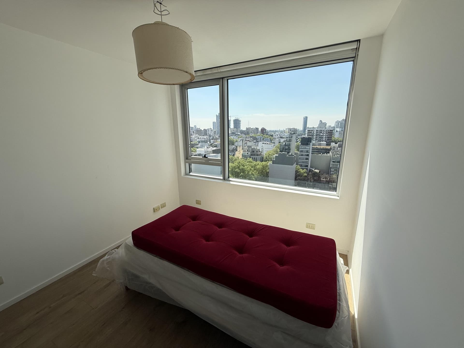 Dormitorio con colchón rojo y ventanal que ofrece vista a la ciudad.