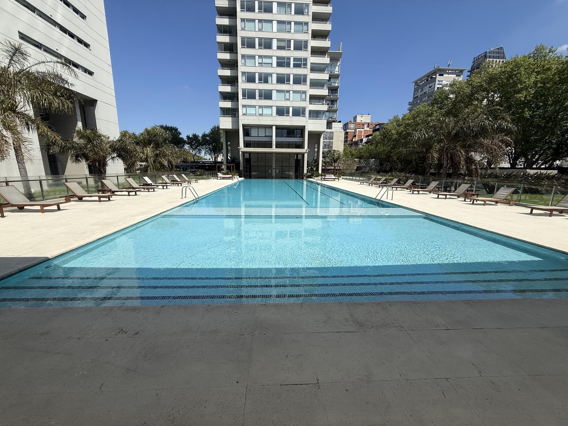 Piscina exterior grande con reposeras y edificios altos de fondo.
