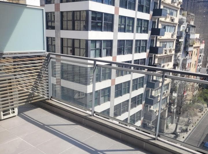 Balcón con piso de baldosas, baranda de vidrio, y edificios altos al fondo.