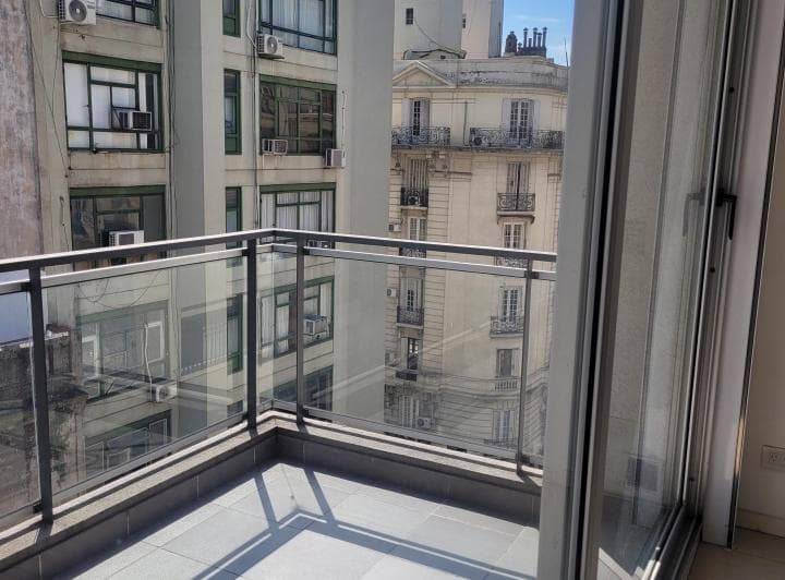 Balcón con piso de baldosas y baranda de vidrio, edificios cercanos.