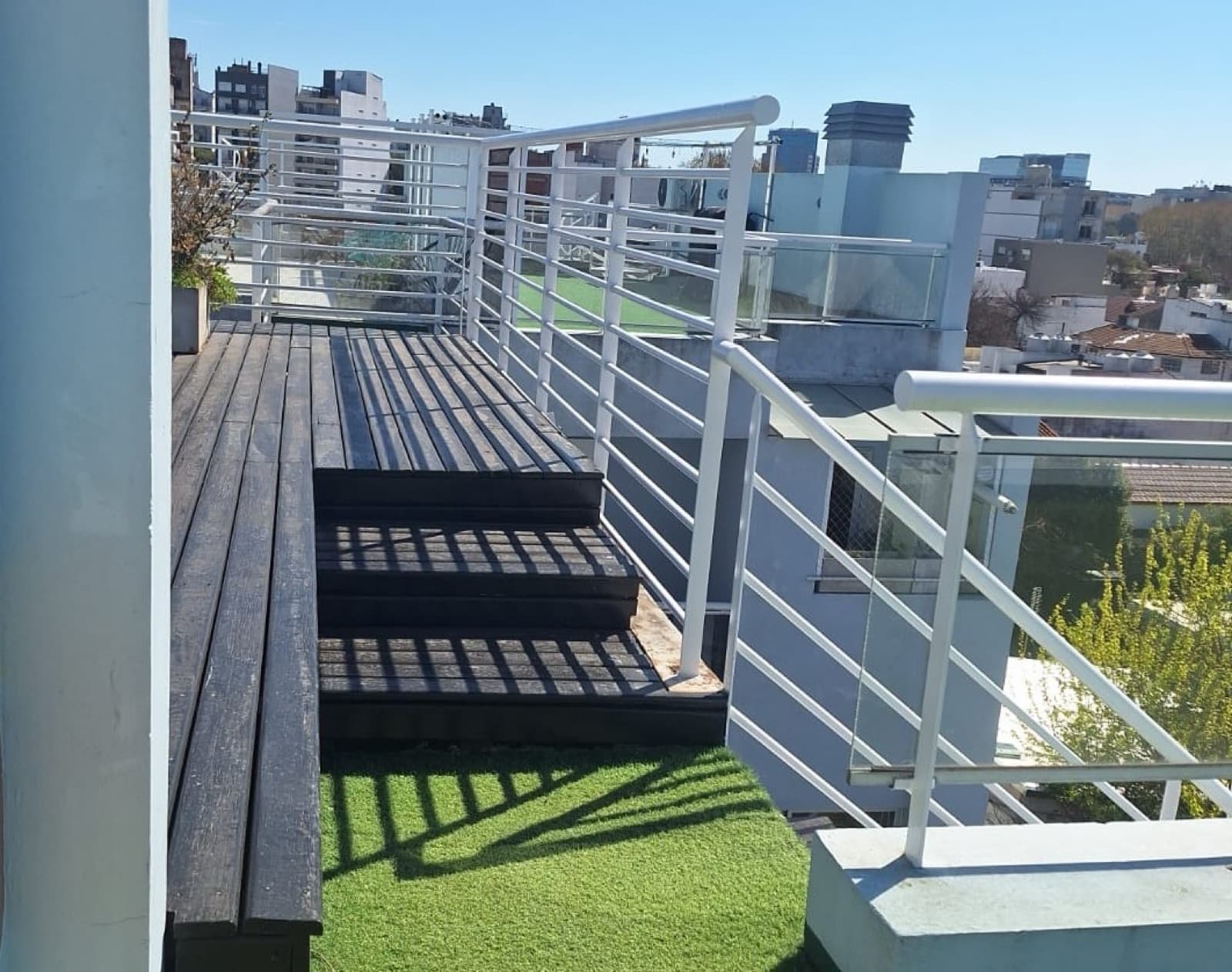 Terraza con escalera de madera, césped artificial y barandas blancas.
