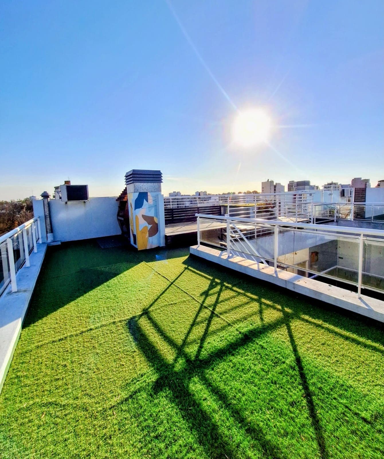 Amplia terraza con césped artificial y vista soleada a la ciudad.
