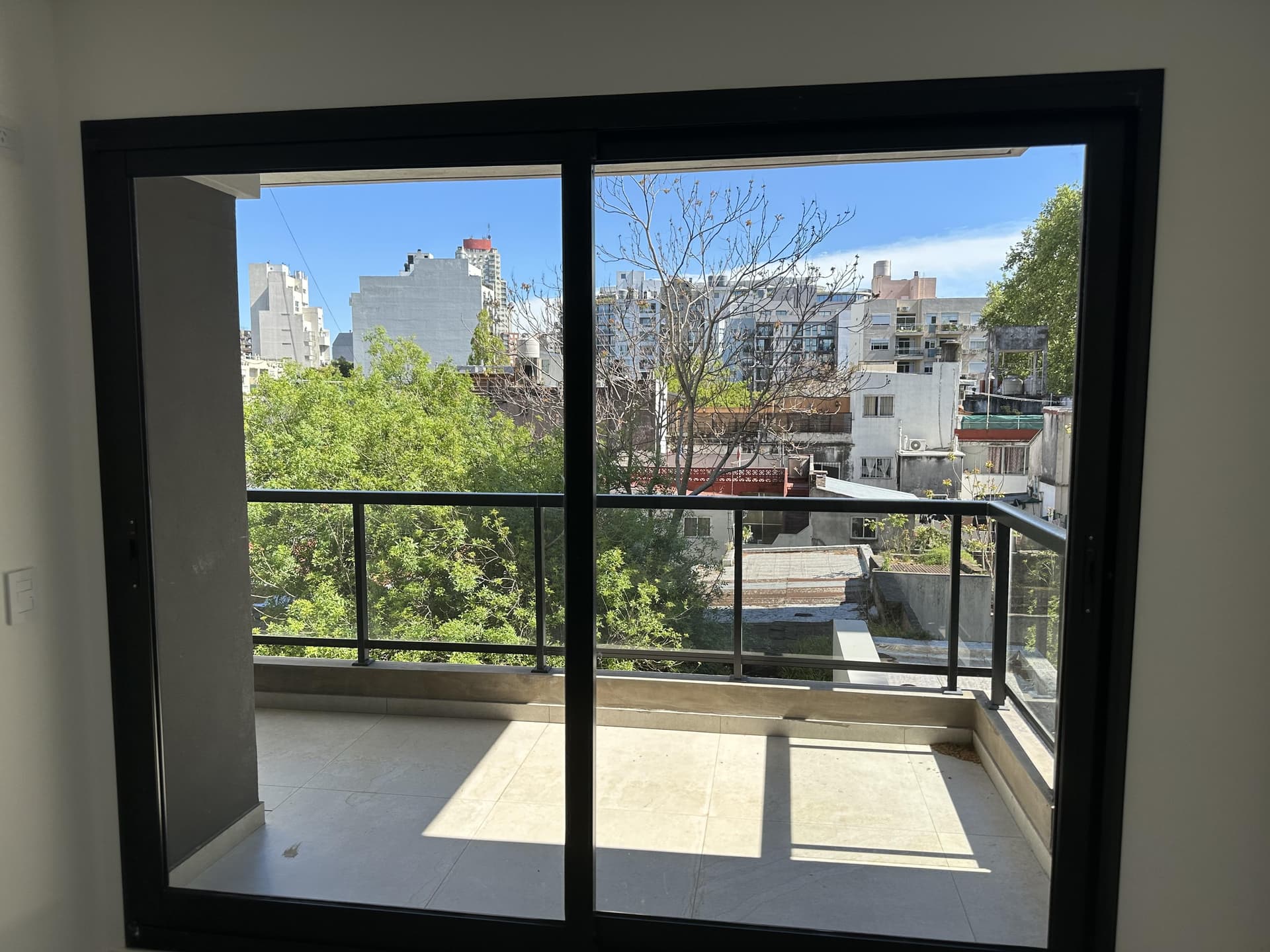Vista desde la ventana hacia un balcón y edificios urbanos.
