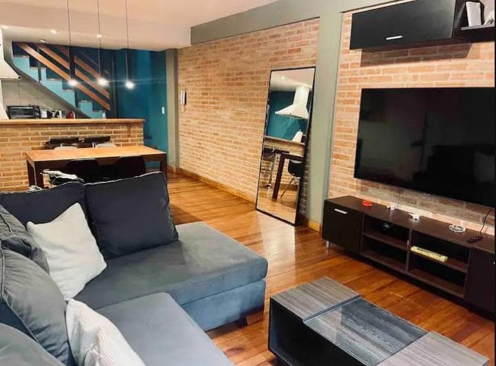 Living con sofá, televisión, pared de ladrillo visto y comedor integrado.