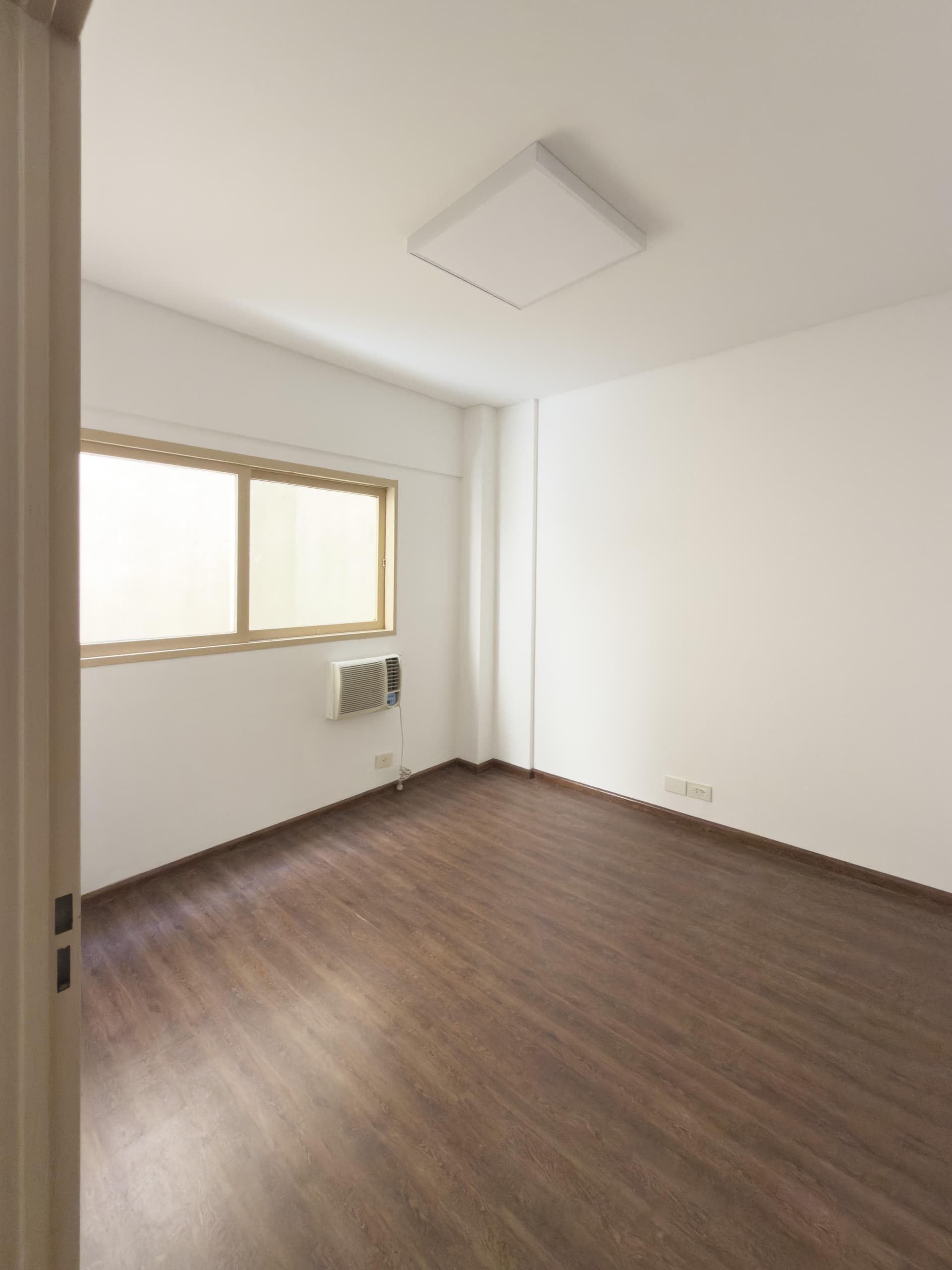 Dormitorio vacío con ventana, aire acondicionado y luminaria de techo cuadrada.