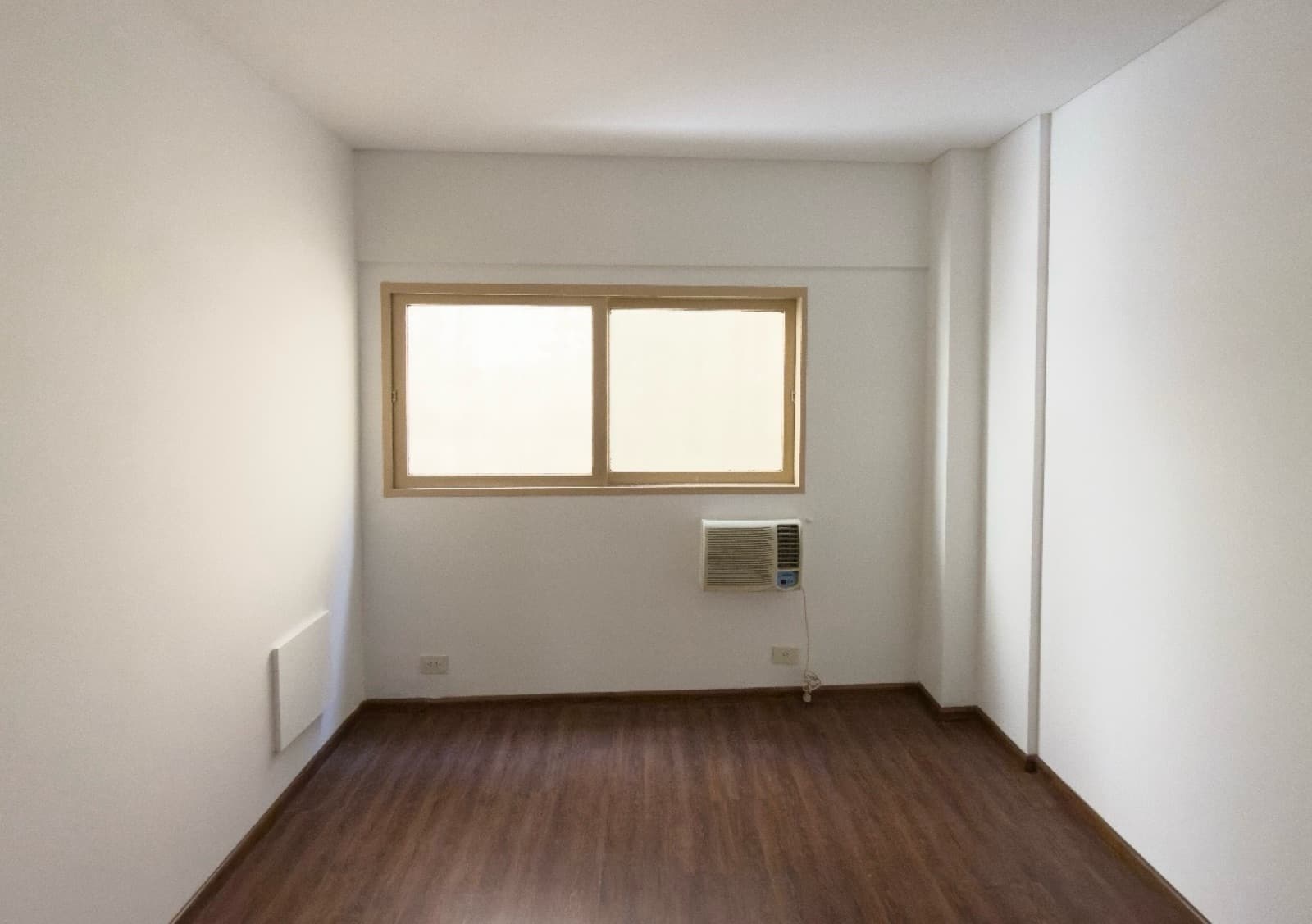 Dormitorio vacío con ventana, aire acondicionado de pared y piso de madera.