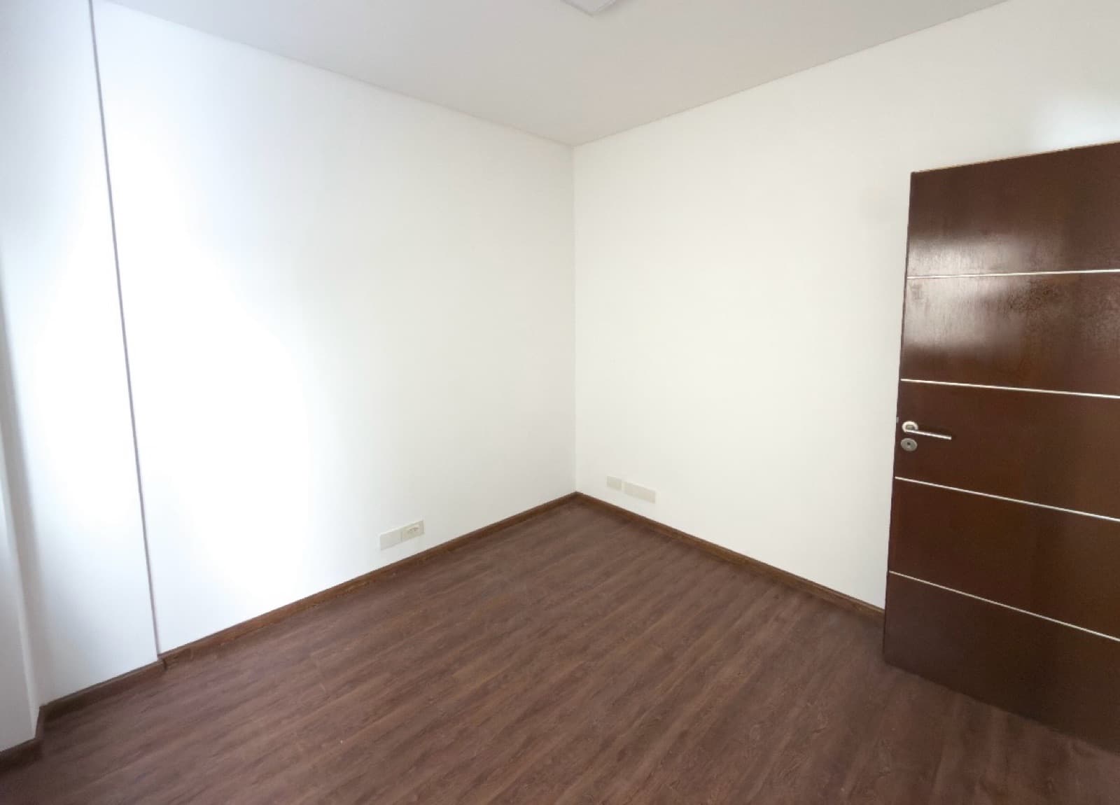 Dormitorio vacío con puerta de madera y piso laminado oscuro.