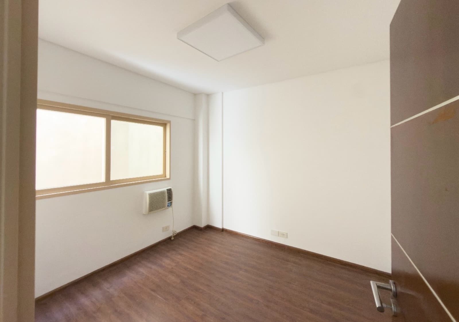 Dormitorio vacío con ventana, aire acondicionado y puerta de madera.