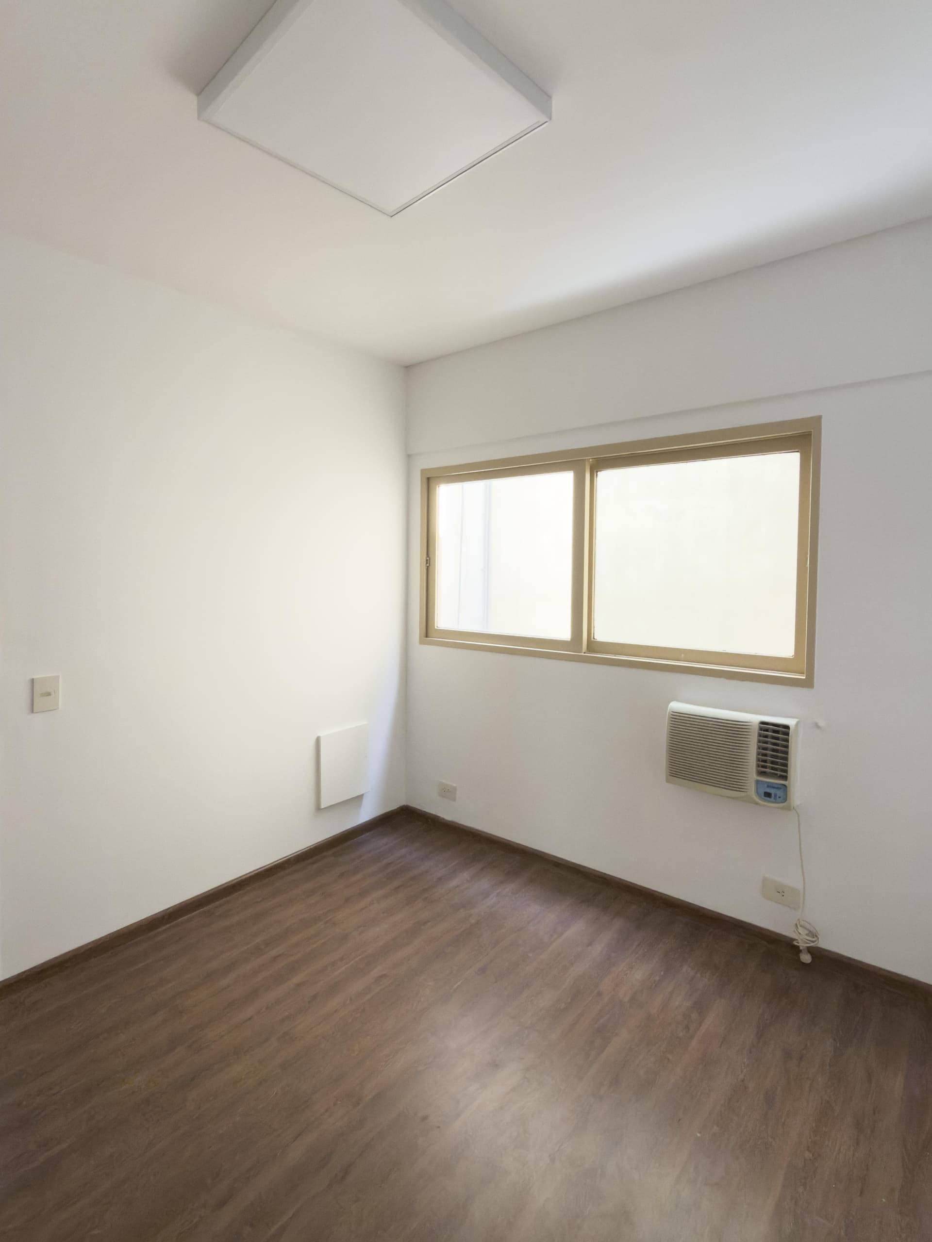 Dormitorio vacío con ventana, aire acondicionado y luminaria de techo cuadrada.