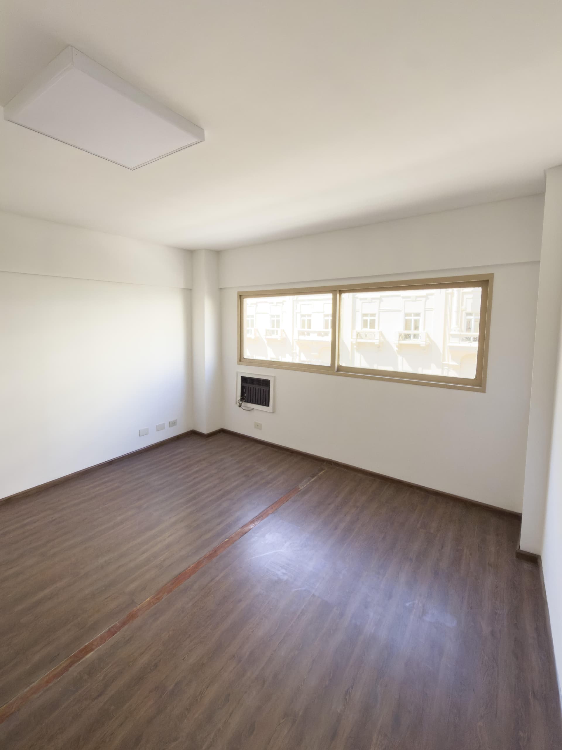 Dormitorio vacío con ventana grande, piso de madera y luminaria.