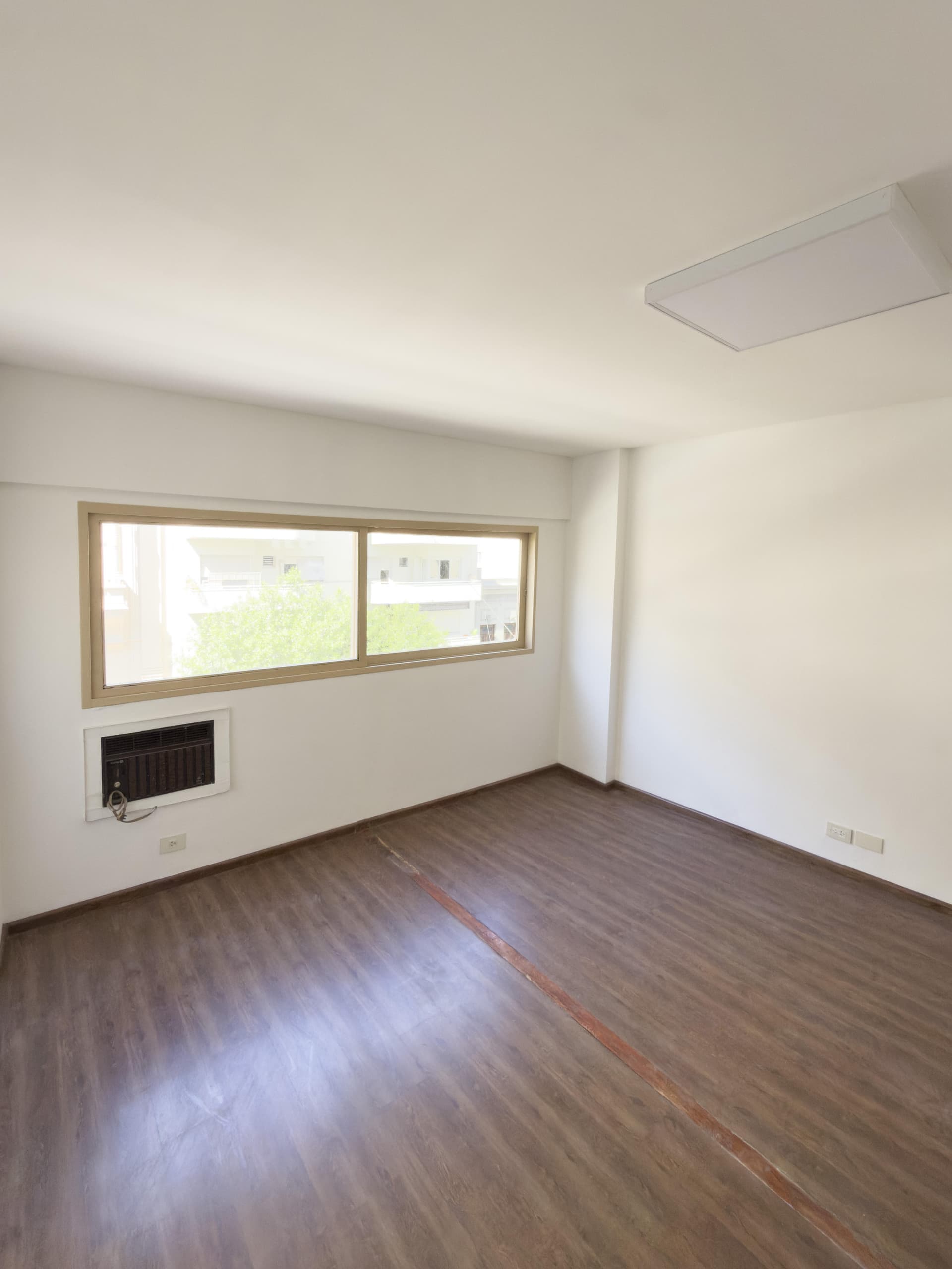 Dormitorio vacío con ventana grande, piso de madera y luminaria.
