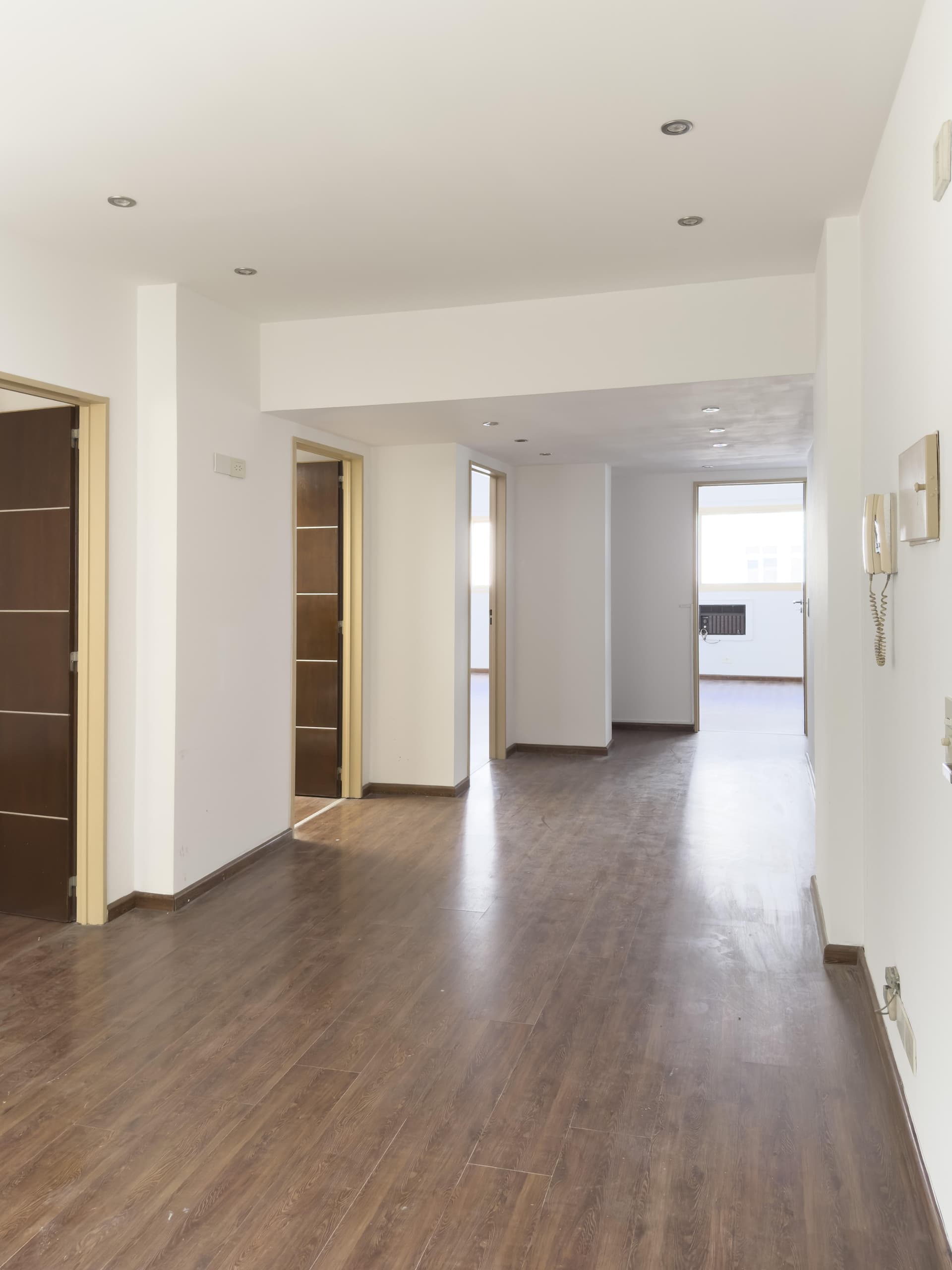 Pasillo largo con piso de madera, paredes blancas y puertas cerradas.