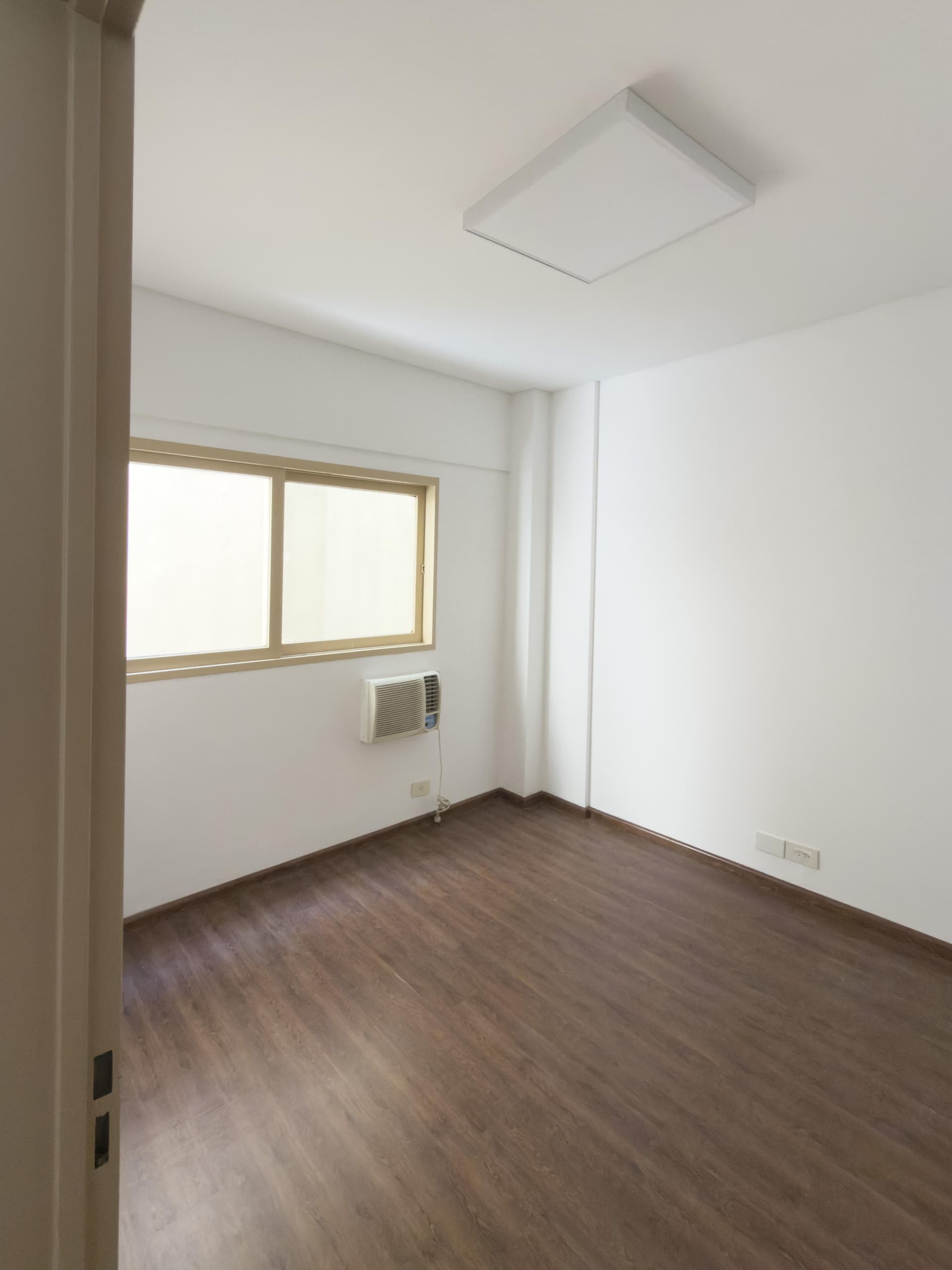 Dormitorio vacío con ventana, aire acondicionado y luminaria de techo cuadrada.