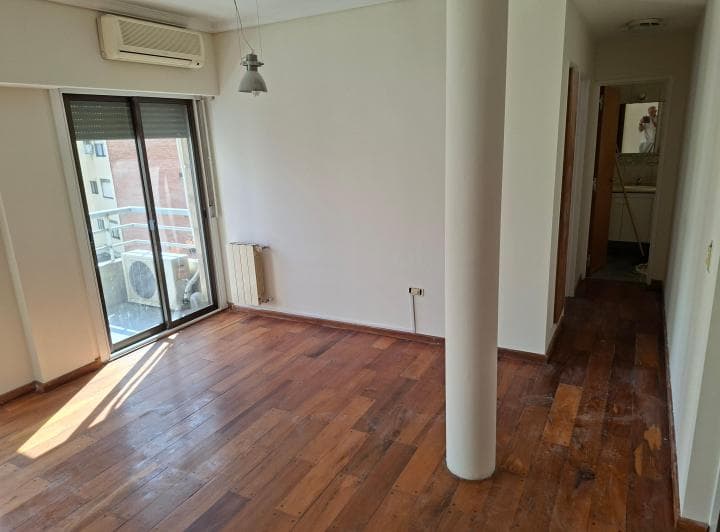 Living vacío con piso de madera, aire acondicionado y acceso a pasillo.