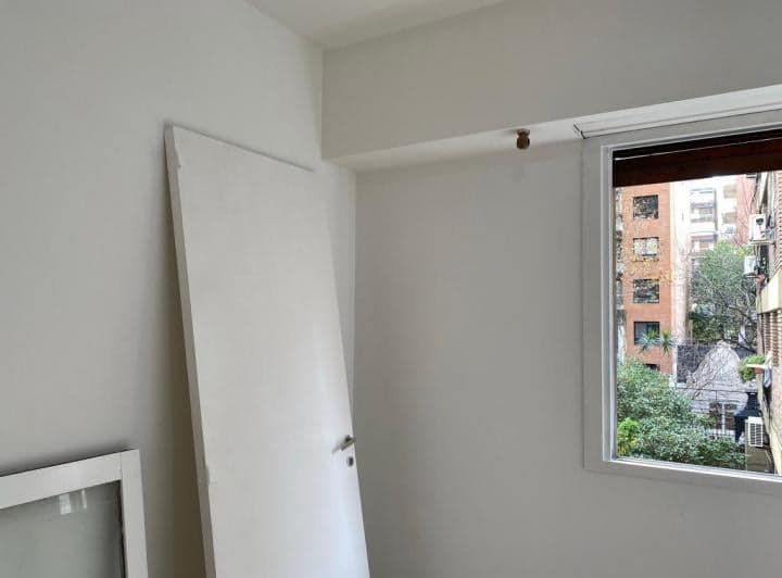 Dormitorio con puerta blanca apoyada en la pared y ventana con vista a edificios.