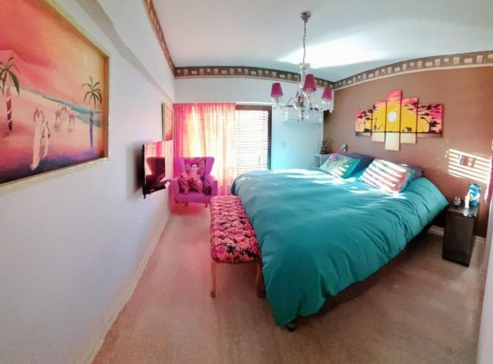 Dormitorio amueblado con cama doble de sábanas azules, cuadros y televisor.