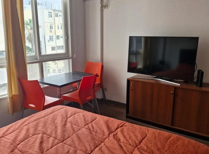 Dormitorio con cama, mesa con sillas y televisor frente a la ventana.