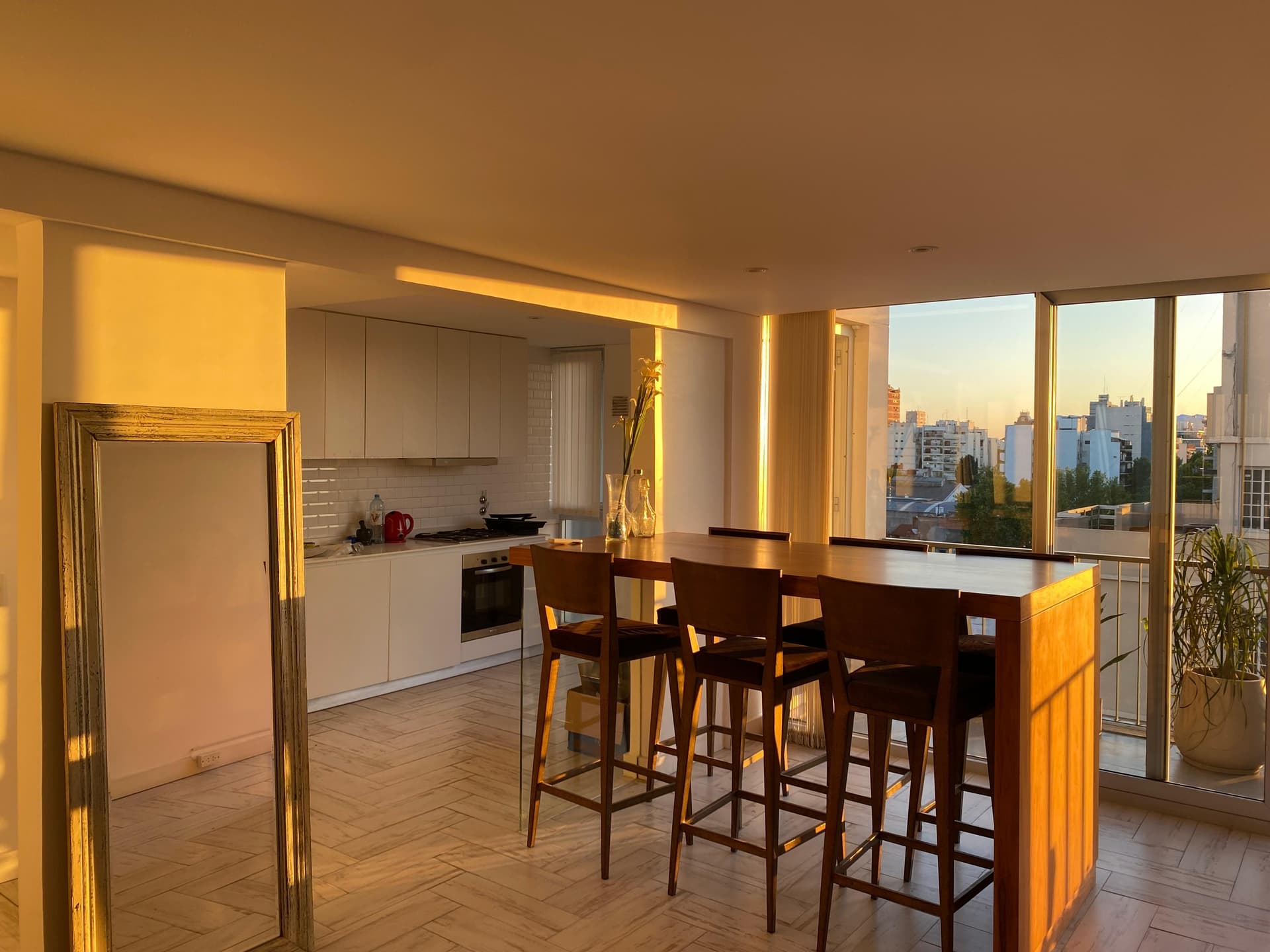 Living con luz del atardecer, mesa alta, cocina integrada y balcón.