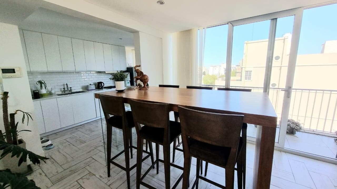 Comedor con mesa alta de madera y banquetas, con cocina integrada al fondo.