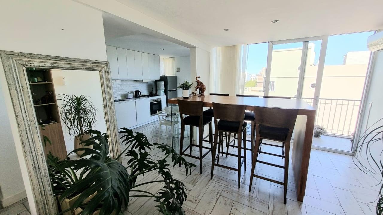 Comedor con espejo de pie, mesa alta con banquetas y cocina integrada.