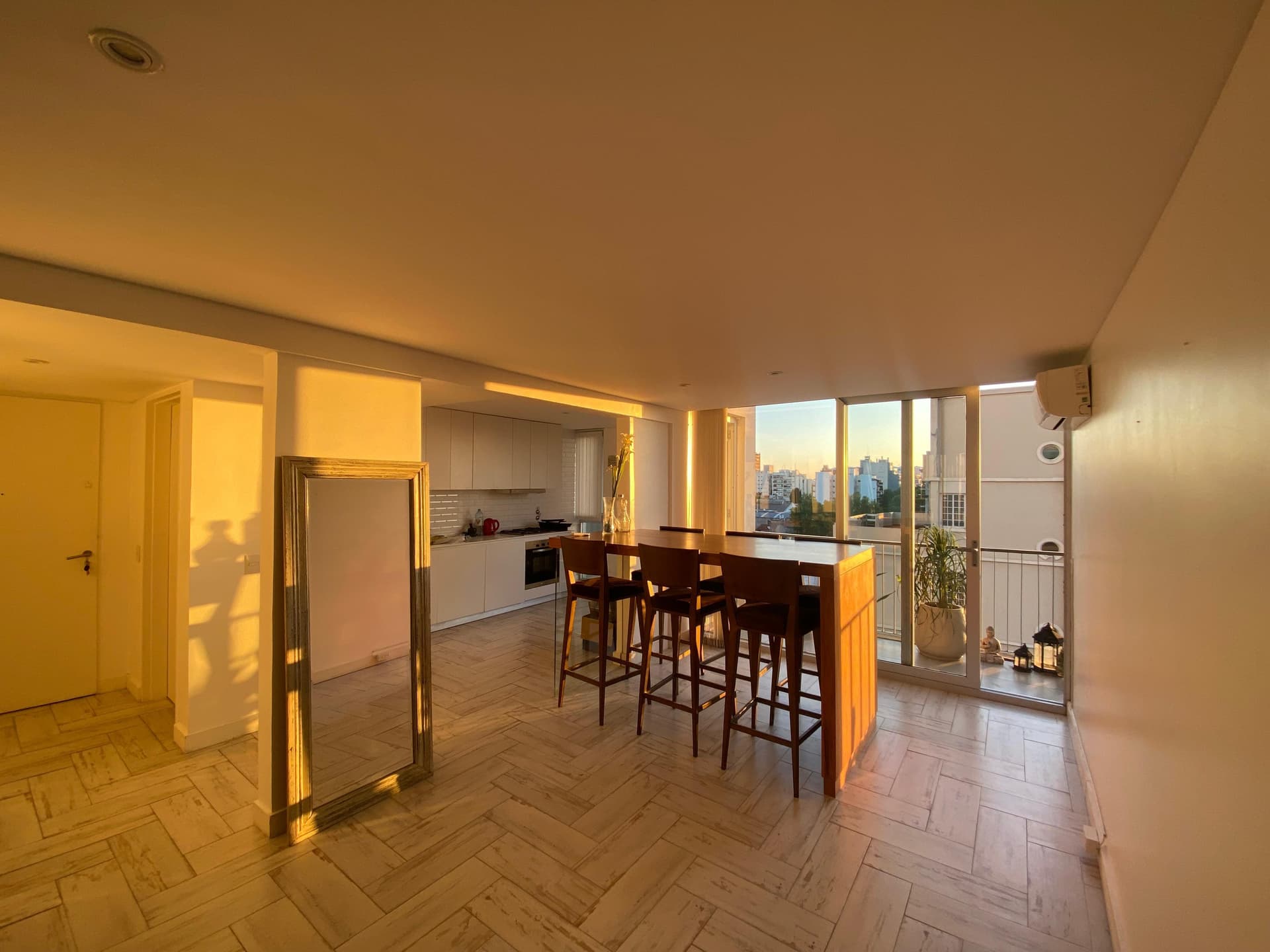 Living con luz del atardecer, mesa alta, cocina integrada y ventanal al balcón.