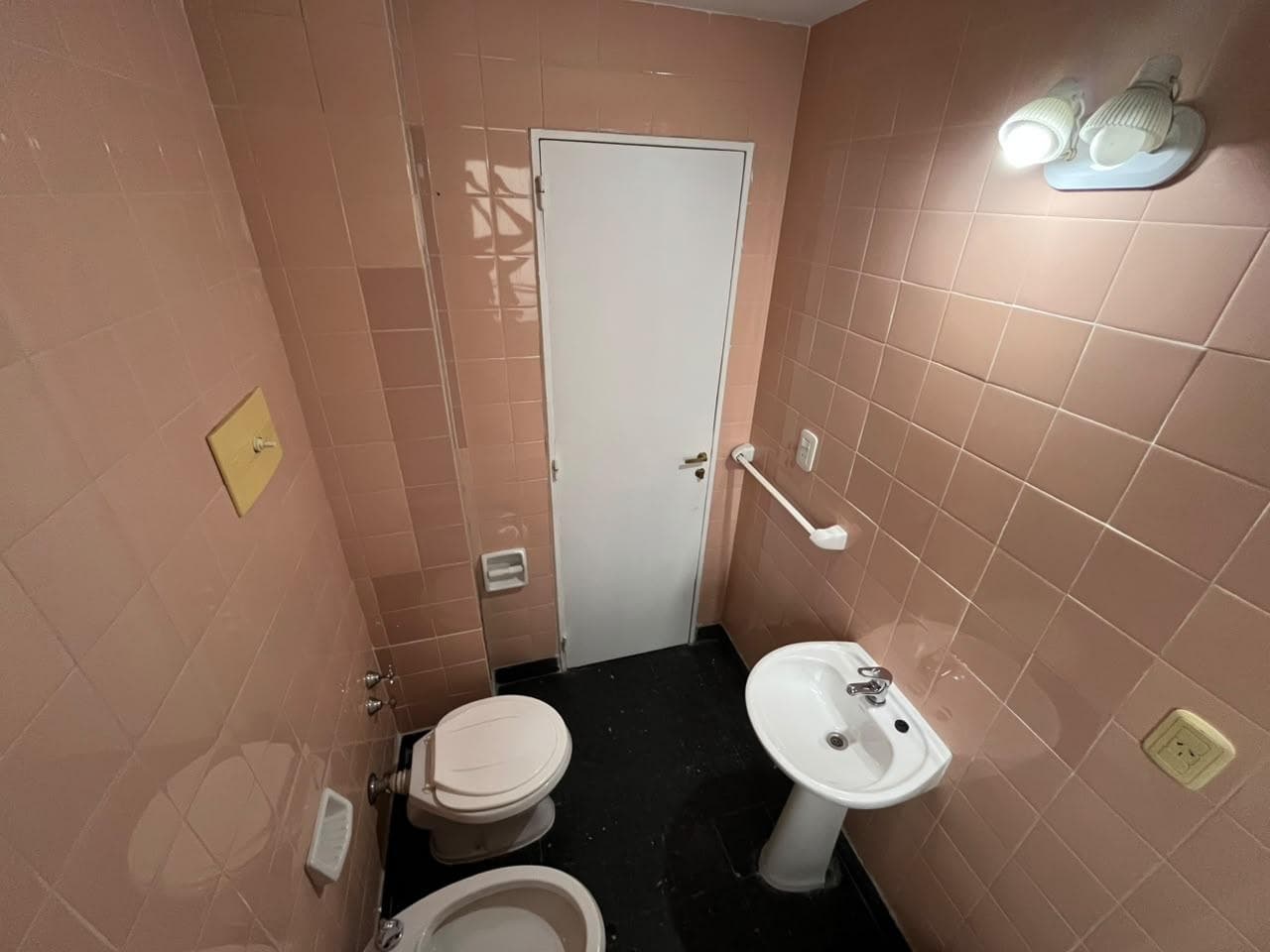 Baño con inodoro, lavatorio y bidet, paredes con azulejos rosas.
