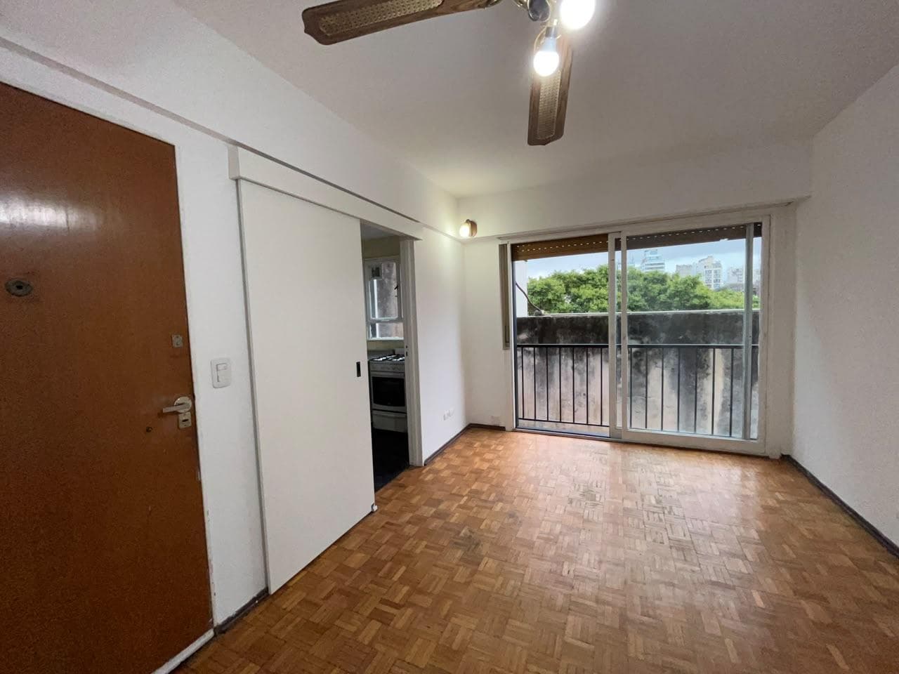Living con piso de parquet, ventilador de techo y puerta a cocina.