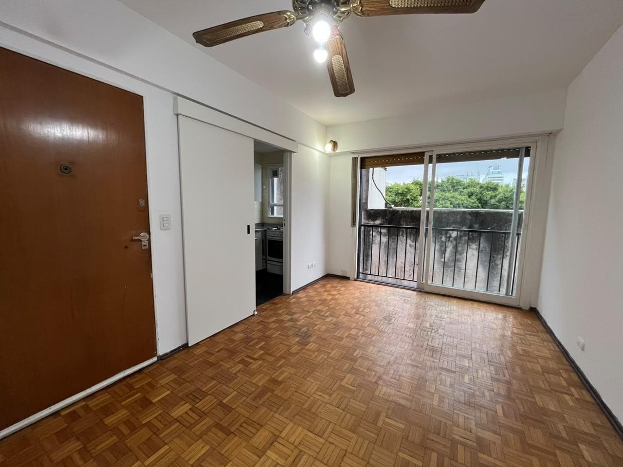 Living con piso de parquet, ventilador de techo y puerta a cocina.