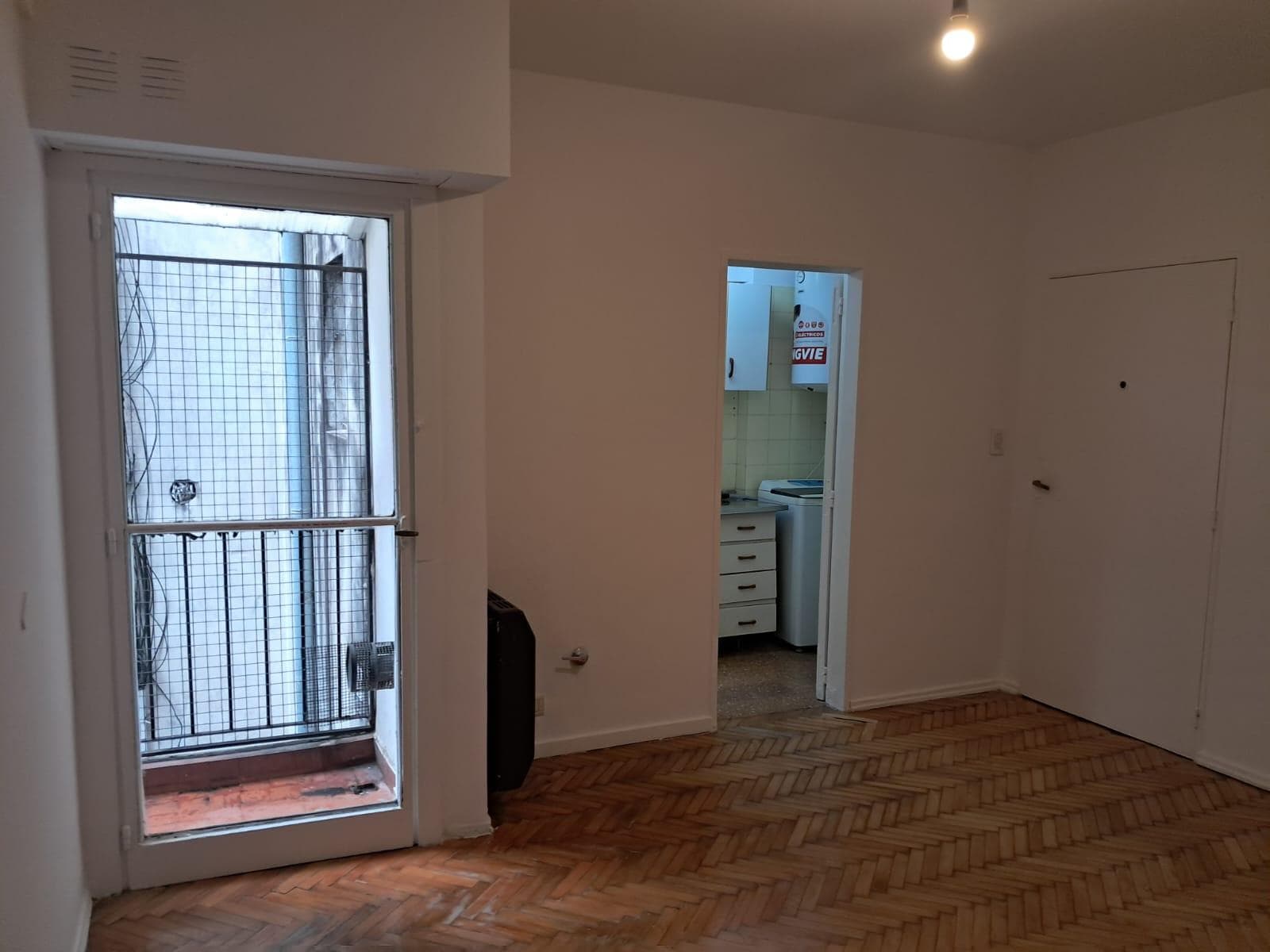 Living vacío con piso de madera, balcón y vista al interior de la cocina.