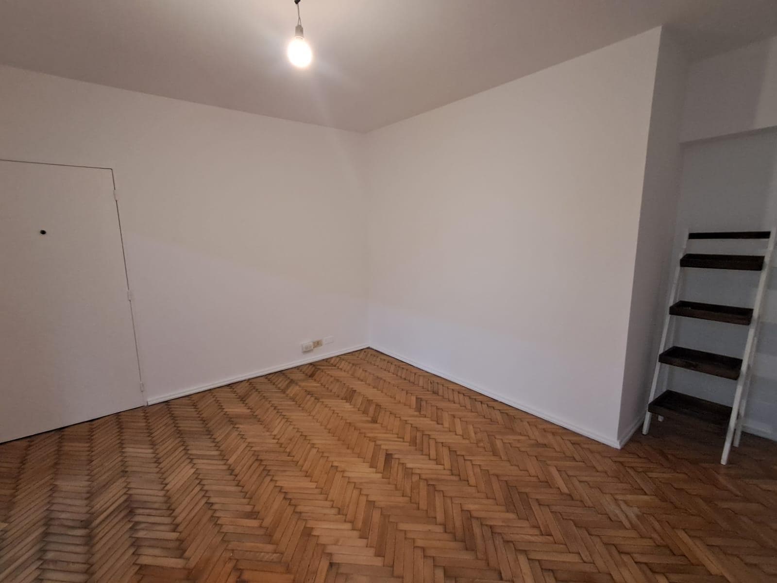 Living vacío con piso de madera en espiga y estantería de madera.