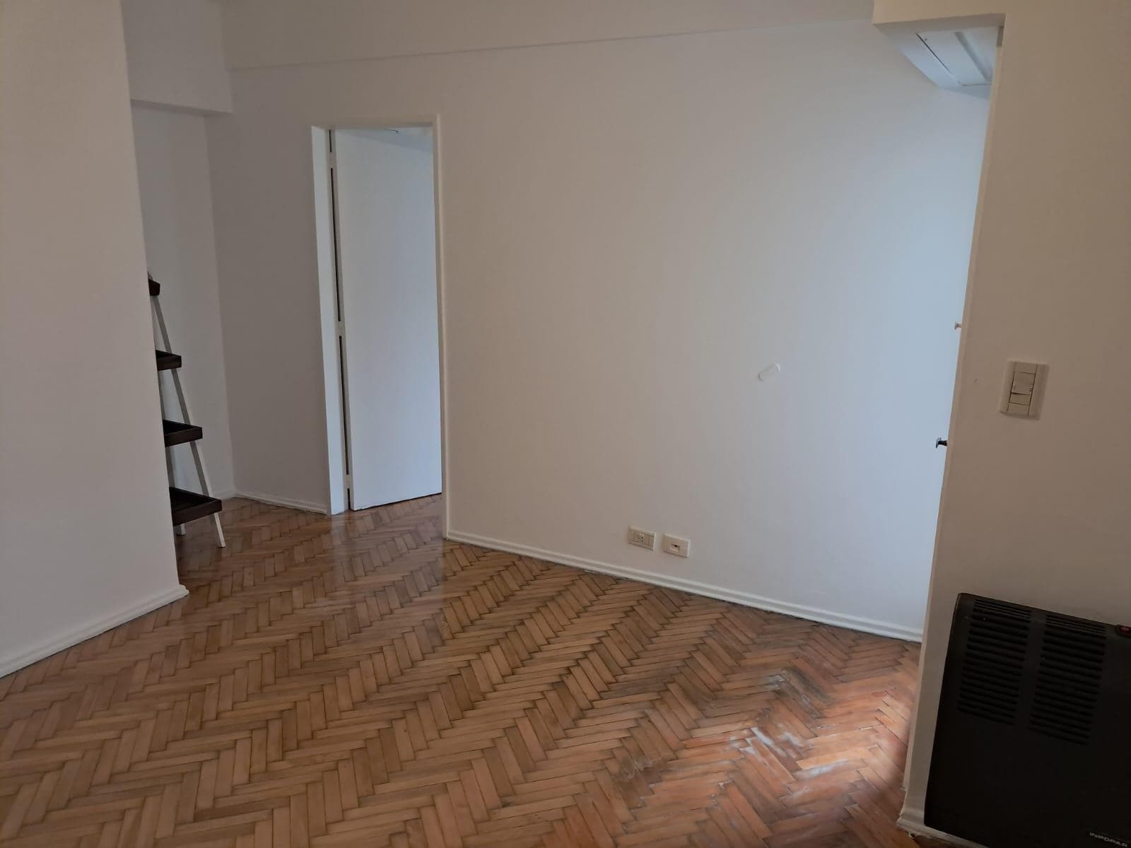 Living vacío con piso de madera, estantería y puerta abierta.