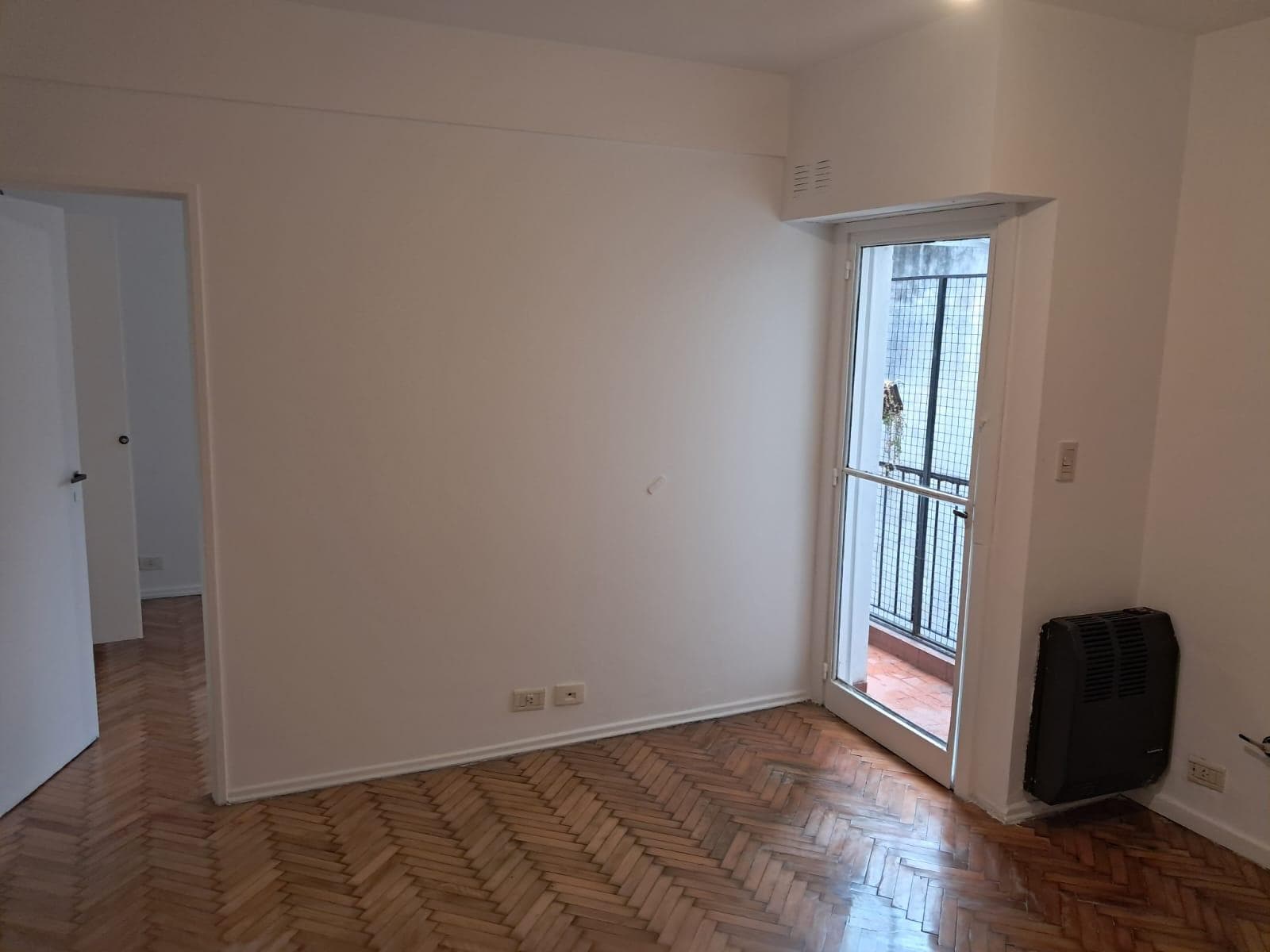 Living con piso de madera, calefactor y salida a un pequeño balcón.