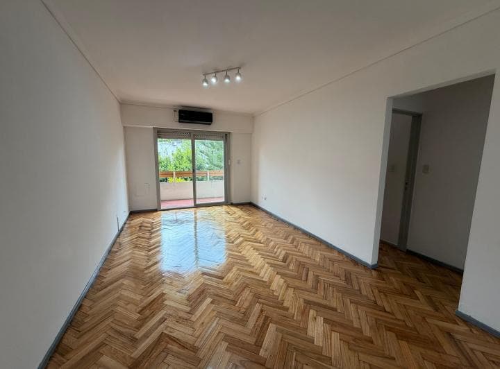 Living vacío con piso de parquet, puerta balcón y aire acondicionado.