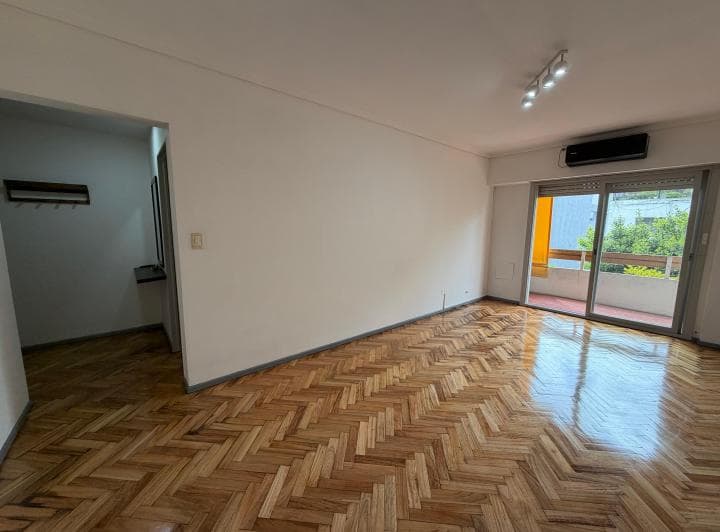 Living con piso de parquet, puerta balcón y acceso a un pasillo.