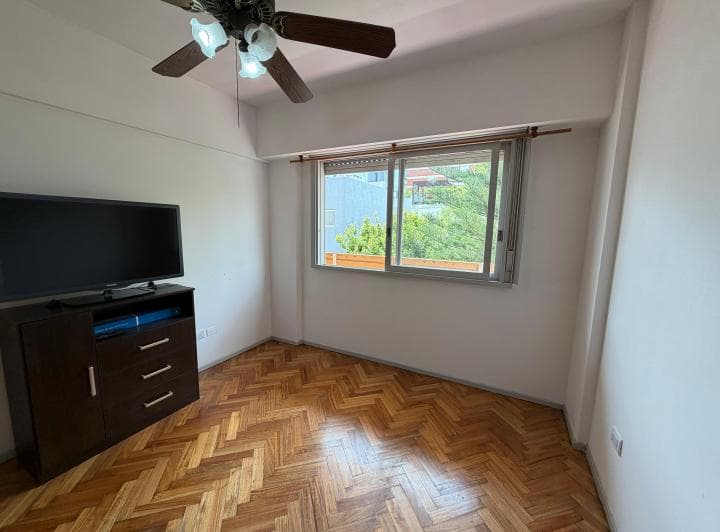 Dormitorio vacío con ventana, piso de parquet y ventilador de techo.