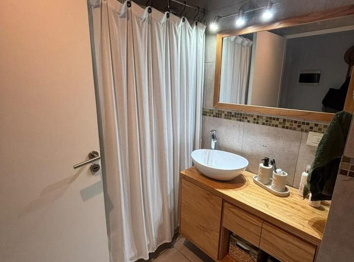 Baño con cortina de ducha, vanitory de madera y bacha de apoyo blanca.