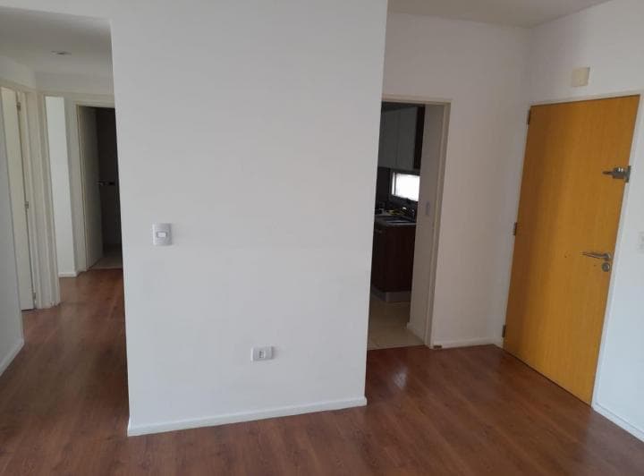 Living con pared blanca, acceso a cocina y pasillo, piso flotante.