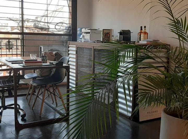 Rincón del living con escritorio, silla, planta y ventana con rejas.