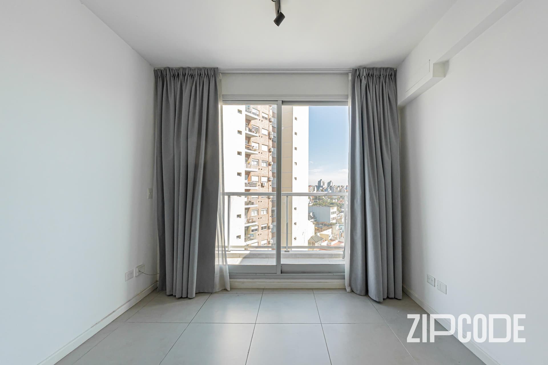 Dormitorio vacío con ventana grande, cortinas grises y vista exterior.